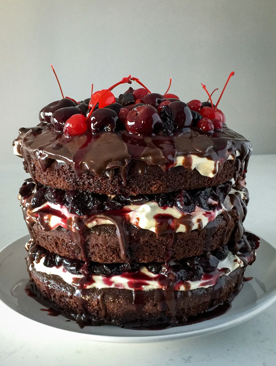 A three-layer chocolate cake piled with cherries on a white stand, in front of the window with the street behind
