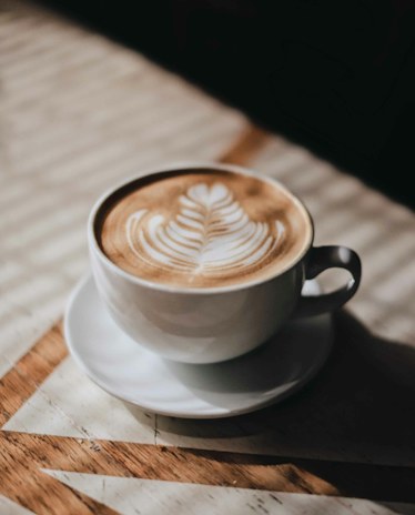 A latte in a white cup on a saucer, sunlight across the wooden table