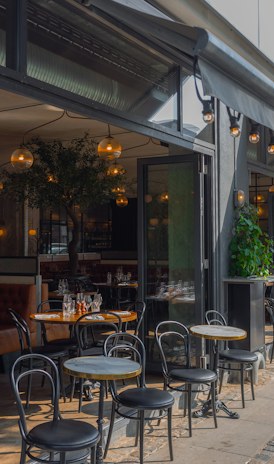 The dark green shopfront with bistro tables and chairs on the pavement and a lamp lit in the window