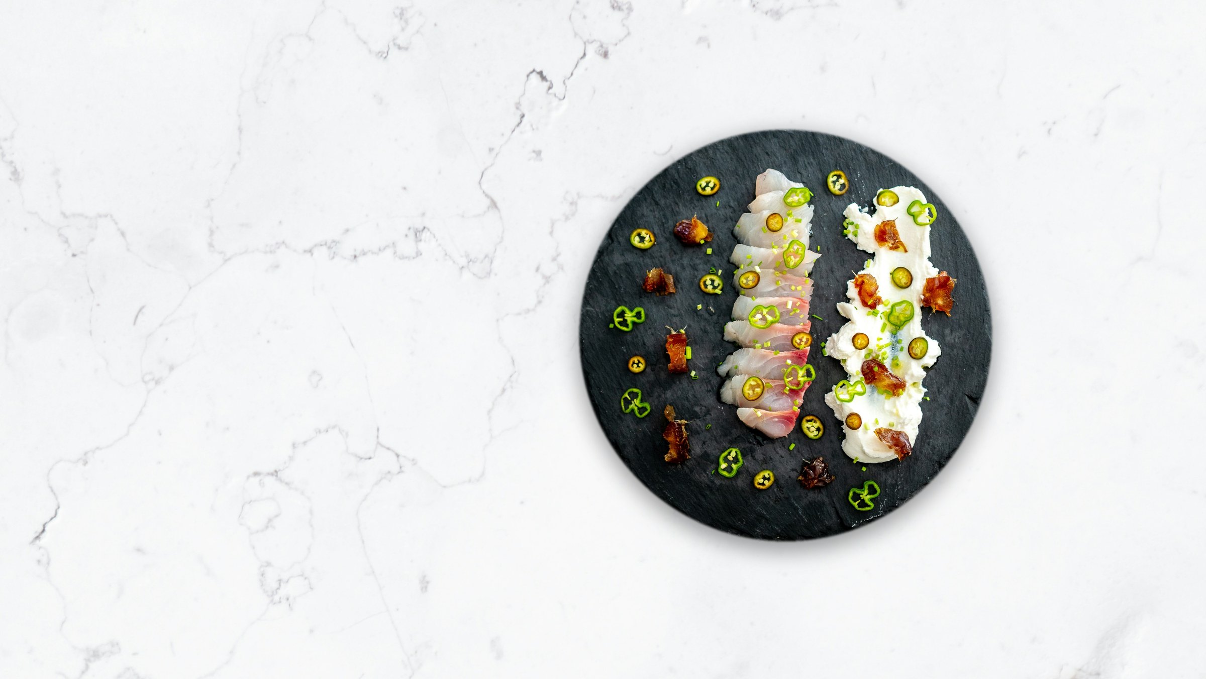 Sliced sashimi and pickles on a round slate plate, on a marble counter.