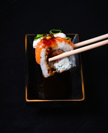 A dynamite roll piece held in chopsticks, topped with shrimp and avocado.